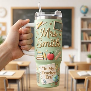 May include: A light green stainless steel tumbler with a handle and straw, featuring raised 3D-style graphics of school supplies, a red apple, a globe, and the text "Mrs. Smith" and "In My Teacher Era" in gold-colored lettering.