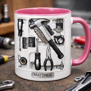 K&ouml;nnte beinhalten: Wei&szlig;e Keramiktasse mit rosa Innenraum und Henkel, mit einem 3D-Werkzeugkasten-Design. Die Tasse enth&auml;lt einen Hammer, Schraubendreher, Zangen und andere Werkzeuge. Der Name "MATTHEW" wird auf einem kleinen Schild angezeigt.