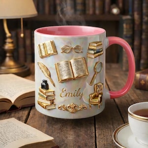 Può includere: Una tazza in ceramica bianca con interno e manico rosa, decorata con abbellimenti dorati a tema libri. La tazza presenta il nome "Emily" in oro ed è appoggiata su una superficie di legno con libri sullo sfondo.