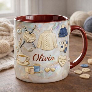 Könnte beinhalten: Eine Keramik-Tasse mit Perlmutt-Finish und bordeauxfarbenem Innenraum und Henkel. Die Tasse ist mit goldfarbenen Verzierungen von Strickartikeln verziert, darunter ein Pullover, Garn, Nadeln und eine Mütze. Der Name "Olivia" steht auf der Tasse.