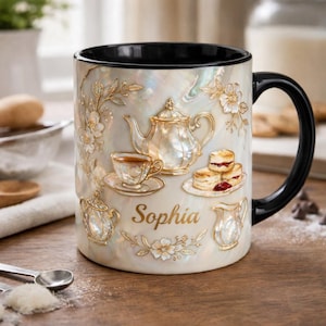 May include: A ceramic mug with a black interior and handle, featuring a pearlescent finish. The mug is adorned with gold-colored embossed images of a teapot, teacup, scones, and the name "Sophia". Floral accents complete the design.