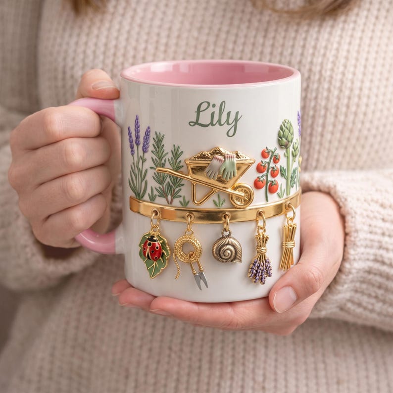 May include: White ceramic mug with a pink interior and handle, decorated with garden-themed illustrations and the name "Lily". The mug features a gold band with hanging charms, including a ladybug, scissors, and a snail. The mug is held in a person's hands.