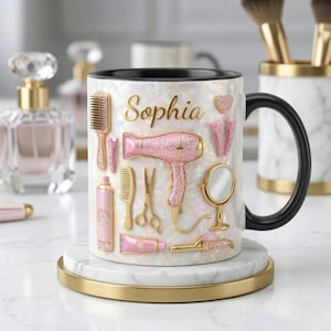 May include: White ceramic mug with a black handle, featuring the name "Sophia" in gold script. The mug is decorated with pink and gold hair styling tools, including a hairdryer, comb, and scissors. The mug sits on a white and gold coaster.