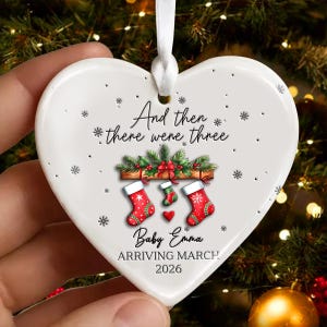 May include: Heart-shaped ceramic Christmas ornament with the text "And then there were three." The ornament features three Christmas stockings, snowflakes, and the text "Baby Emma Arriving March 2026."