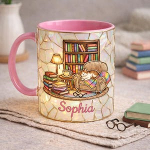 May include: A decorative mug with a pink handle and rim, featuring a reading nook scene with a cat, books, and a lamp. The mug has a pearlescent finish and the name "Sophia".