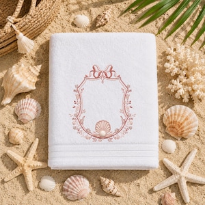 May include: White beach towel with a decorative embroidered frame in shades of pink and a seashell design. The towel is surrounded by seashells, starfish, and a straw bag on a sandy surface.