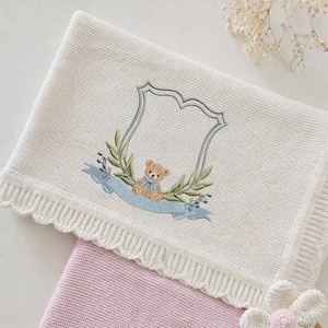 Può includere: Coperta per bambini color crema con un design ricamato. Il design presenta un orsacchiotto, un nastro blu e una corona d'alloro all'interno di una cornice a forma di scudo. La coperta ha un bordo smerlato.