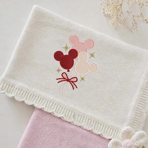 May include: Cream-colored knitted baby blanket with a scalloped edge. The blanket features an embroidered design of three Mickey Mouse head-shaped balloons in red, pink, and beige, with gold star accents and a red bow.