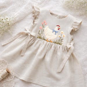 May include: A light beige linen dress with an embroidered hen design. The dress features a hen with a blue bow, pink flowers, and daisies. The dress has ruffled sleeves and tie closures, suitable for a young girl.