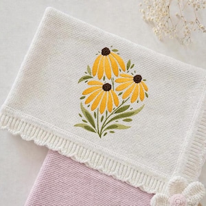 May include: A white knitted blanket with a yellow flower embroidery design. The blanket has a scalloped edge and is folded over a pink blanket. The embroidered flowers have brown centers and green leaves.