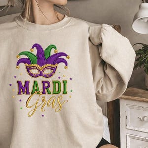 Puede incluir: Sudadera beige con un diseño de Mardi Gras. El diseño presenta un sombrero y una máscara de bufón morados y verdes, con las palabras "Mardi Gras" en letras doradas y verdes. Estrellas doradas están dispersas alrededor del texto.