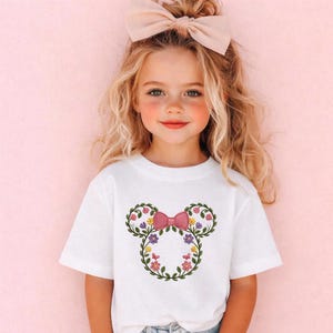 May include: White t-shirt featuring a floral Mickey Mouse design. The design includes a pink bow, green leaves, and colorful flowers. The girl in the image is wearing a matching pink bow in her hair.