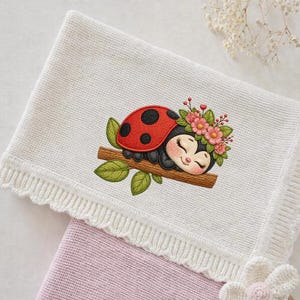 May include: A white knitted baby blanket with a scalloped edge and a ladybird design. The ladybird is red with black spots, resting on a branch with green leaves and pink flowers. A second pink blanket is visible underneath.