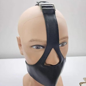 Puede incluir: Una máscara facial de material similar al cuero negro texturizado con una correa vertical que se extiende sobre la nariz y la frente, asegurada con una hebilla de metal plateado en la parte superior. La máscara cubre la parte inferior.