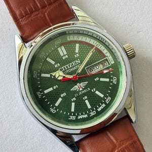 May include: A Citizen automatic watch with a green dial, silver case, and brown leather strap. The watch face displays the day and date, with white numerals and hands. The watch reads "21 JEWELS" and "JAPAN MADE".