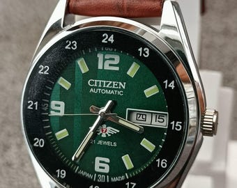 May include: A Citizen Automatic watch with a green face and a brown leather band. The watch face displays the time with white numerals and markers, and the date. The watch has the text "CITIZEN AUTOMATIC" and "21 JEWELS".