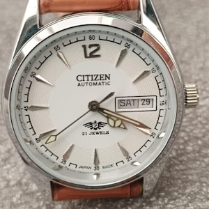 Könnte beinhalten: Eine silberne Citizen Automatik-Armbanduhr mit weißem Zifferblatt, schwarzen Ziffern und Zeigern. Die Uhr hat ein braunes Lederarmband und zeigt Tag und Datum an. Auf dem Zifferblatt steht "CITIZEN AUTOMATIC" und "21 JEWELS".