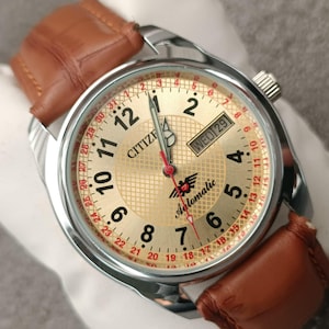 May include: A Citizen automatic wristwatch with a gold-colored face, silver casing, and a brown leather strap. The watch face displays the day and date, with black numerals and red accents. The watch is a classic timepiece.