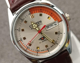 Puede incluir: Reloj automático Citizen plateado con correa de cuero marrón. La esfera del reloj presenta detalles en naranja y negro, agujas doradas y una pantalla de día y fecha. El reloj está marcado "21 JEWEL" y "JAPAN".