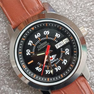Puede incluir: Un reloj Citizen Automatic con esfera negra, bisel plateado y correa de cuero marrón. La esfera muestra el día y la fecha, con números blancos y detalles en naranja. Las manecillas son rojas. El reloj está ajustado al viernes 21.
