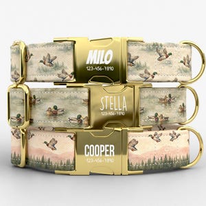 May include: Three personalized dog collars with gold-colored buckles. The collars feature a duck and landscape pattern. Each buckle is engraved with a name and phone number: MILO, STELLA, and COOPER.