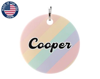 May include: A round, pastel-colored pet tag with diagonal stripes in pink, yellow, green, and purple. The name "Cooper" is written in black script across the center. A silver-colored ring is attached at the top. The tag has a "Made in the USA" sticker.