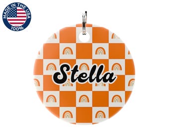 May include: A round, orange and white checkered tag with the name "Stella" in black script. The tag features rainbow graphics and a silver keyring. The tag is made in the USA.