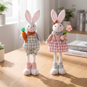 May include: Two Easter bunny figurines. One bunny wears a green and white checkered overall with a carrot. The other bunny wears a pink and white checkered dress and holds a pink flower. Both have long white legs and large ears.