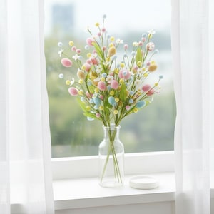 May include: A colorful floral arrangement in a clear glass vase. The bouquet features pastel-colored egg-shaped ornaments, small white flowers, and green leaves. The vase sits on a white windowsill near a sheer white curtain.