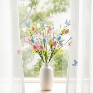 May include: A white ceramic vase holds a spring floral arrangement. The arrangement features pastel-colored eggs, butterflies, and bunny figures. The flowers are pink, blue, and yellow, with green stems. The background shows a window with sheer white curtains.