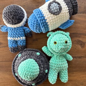 May include: A collection of handmade crochet space-themed toys. Includes a blue and white rocket, an astronaut in a blue suit, a teal alien with yellow eyes, and a gray and teal UFO. The toys are made of soft, textured yarn.