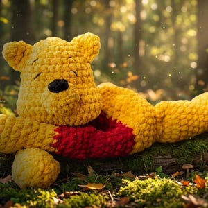 May include: A yellow crocheted bear plush toy with a red shirt and a black nose. The bear is lying down with its head resting on its arm, featuring a textured yarn construction with visible stitches throughout the entire body of the figure.