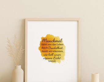 May include: A framed print with a light wood frame. The print features the German text "Manchmal" in a script font, with a yellow and orange watercolor design. A small vase with dried plants is to the left.