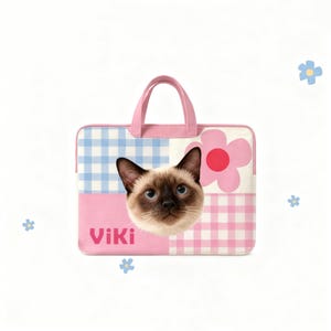 May include: A pink and blue laptop case with a Siamese cat face. The case has a pink handle and a flower design. The name "Viki" is printed on the front. The case has a gingham pattern.