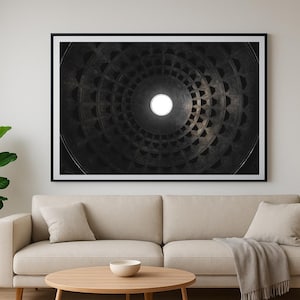 Puede incluir: Una impresión en blanco y negro enmarcada de un techo arquitectónico circular con una fuente de luz central. La obra de arte cuelga sobre un sofá beige con cojines y una manta. Una planta en maceta y una lámpara de pie negra también están en la habitación.