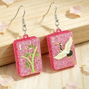 Puede incluir: Un par de pendientes rosas con purpurina y ganchos plateados. Un pendiente presenta un diseño de bambú y el número 1, el otro tiene una grulla y el número 2. Los pendientes son rectangulares y tienen un fondo rosa brillante.