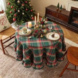 Puede incluir: Un mantel redondo con un patrón de cuadros verdes, rojos y amarillos con un borde con volantes. El mantel está dispuesto con platos blancos, una taza floral, dos velas en candelabros dorados, un centro de mesa de piñas y un vaso.