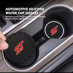 May include: Two round black automotive silicone water cup gaskets featuring a red and white ST logo in the centre. The coasters have a textured outer rim and are designed to fit into a vehicle cup holder to prevent noise while driving.