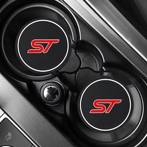 May include: Black car cup holder coasters with a white border and the red letters "ST" in the centre. The coasters are circular and designed to fit inside a car's cup holders. The car's interior is black.