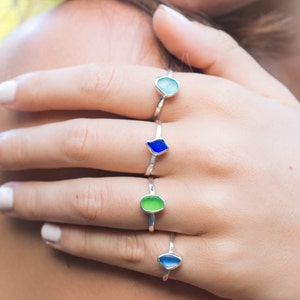 Puede incluir: Una mano con cuatro anillos de plata con piedras de vidrio de diferentes colores. Los anillos están apilados en el dedo. Las piedras son azules, verdes y azul claro.
