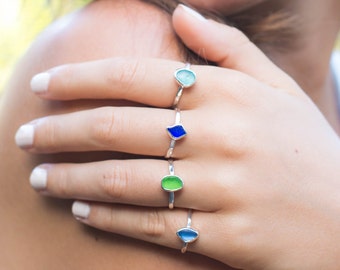 May include: A hand wearing four silver rings with different coloured glass stones. The rings are stacked on the finger. The stones are blue, green, and light blue.