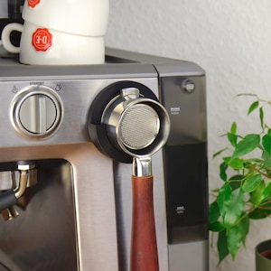 Puede incluir: Máquina de espresso de acero inoxidable con un portafiltro con mango de madera. Dos tazas de café blancas con logotipos rojos están apiladas. A la derecha hay una planta verde y frondosa en maceta.