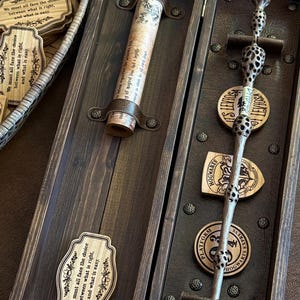 Puede incluir: Una caja de madera oscura que contiene una varita, un pergamino y placas decorativas de madera. La varita es plateada con un mango detallado. El pergamino está sujeto por correas de cuero. Las placas tienen texto y diseños florales.