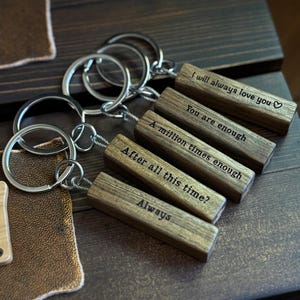 Peut inclure: Porte-clés en bois avec des anneaux argentés. Chaque porte-clés est composé d'un bloc de bois rectangulaire gravé de textes : "(I will always love you)", "You are enough", "A million times enough", "After all this time?", et "Always".