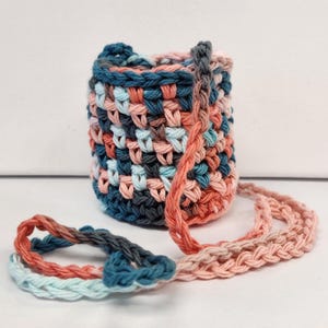 May include: A small, handmade crochet bag in a mix of colors including coral, teal, and gray. The bag has a woven texture and a long, braided strap. The bag is approximately 5 inches tall and 4 inches wide.