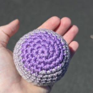 May include: A crocheted ball in shades of purple and gray, held in a hand. The ball's texture is visible, with a spiral pattern in the center. The color transitions from a deep purple to a lighter gray.