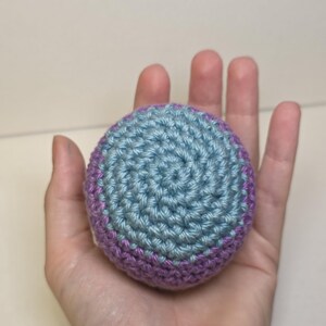 May include: A spherical crochet object featuring a central circular pattern in light blue yarn, surrounded by a border of purple yarn. The item is constructed with a tight, consistent stitch pattern, creating a textured, rounded surface.