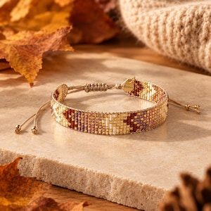 Op de afbeelding: Een geweven kralenarmband met een geometrisch patroon in goud-, bruin-, crème- en mauvetinten. De armband heeft een verstelbare koordsluiting met twee kleine goudkleurige kralen aan de uiteinden en een klein goudkleurig label met de tekst Bystyn.