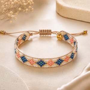 Könnte beinhalten: Ein gewebtes Perlenarmband mit einem sich wiederholenden Rautenmuster in Blau-, Rosa- und Weißtönen. Das Armband hat einen verstellbaren Kordelverschluss mit goldfarbenen Perlen an den Enden und einen kleinen runden, goldfarbenen Metallanhänger mit ByStyn-Gravur.