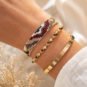 Op de afbeelding: Een set van drie armbanden bestaande uit een brede geweven kraalarmband met een geometrisch patroon in bordeauxrood, crème en metallic goud, een fijne goudkleurige kettingarmband met kleine ovale schakels en een gladde goudkleurige armband.